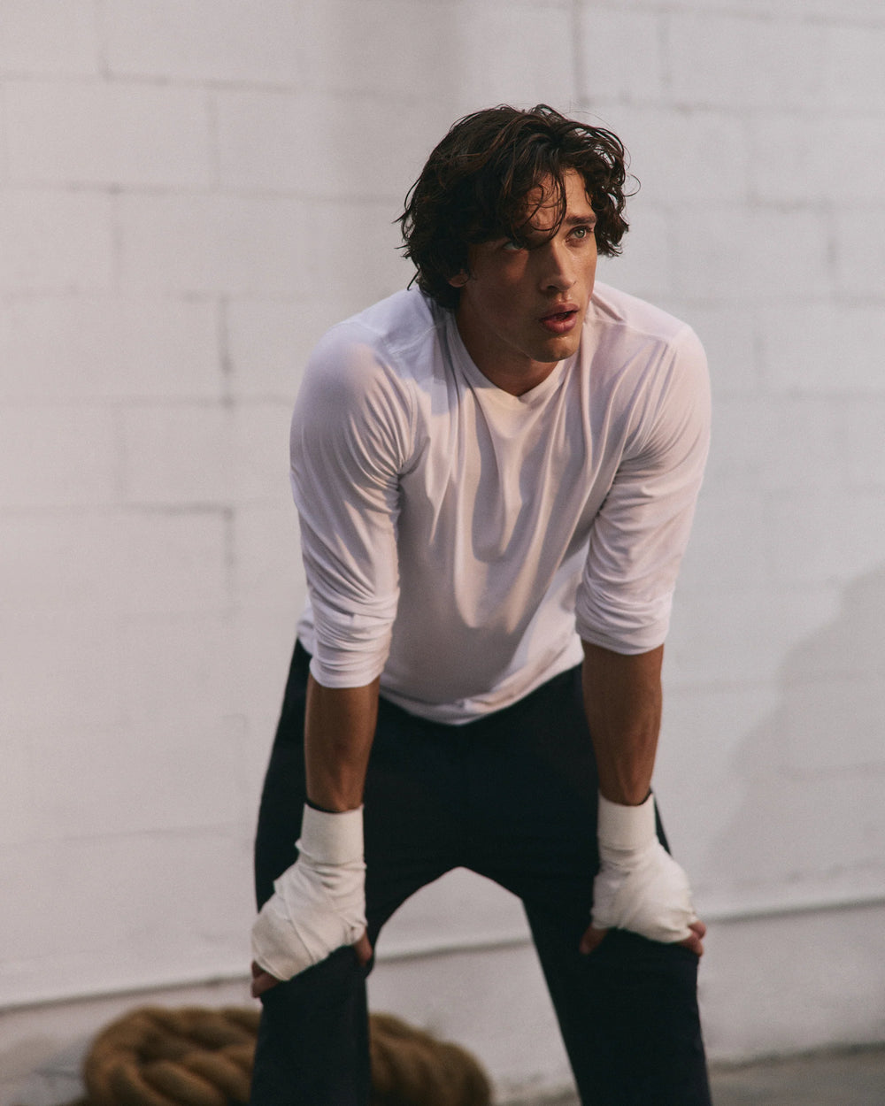 Person wearing a white long-sleeve shirt and black pants with white boxing gloves against a light gray wall.