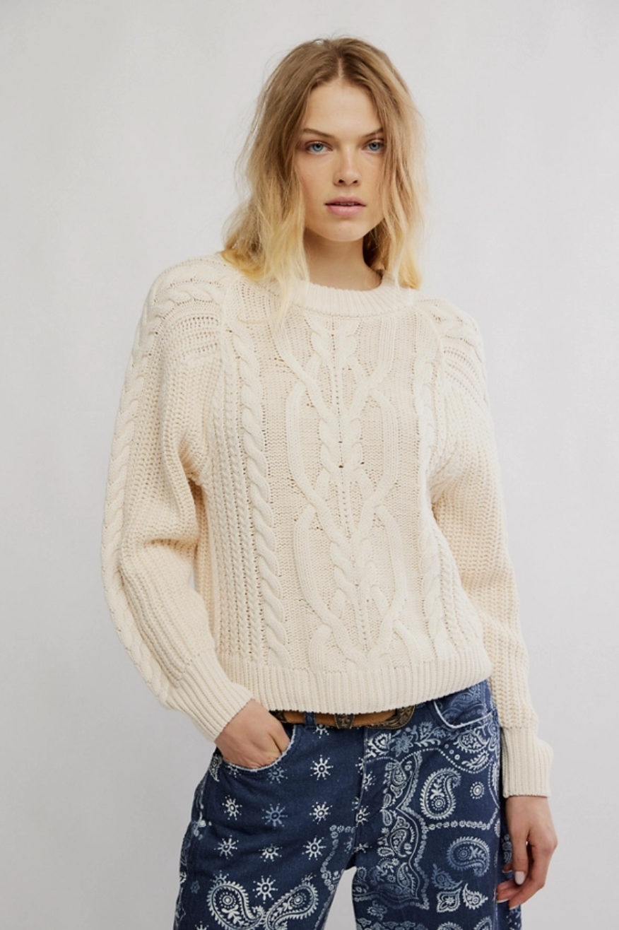 Woman wearing a cream cable knit sweater and blue patterned pants on a plain background