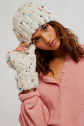 A woman wearing a chunky knit beanie and matching fingerless gloves in an off-white yarn with multicolored flecks, paired with a dusty pink henley top, against a plain light gray background.