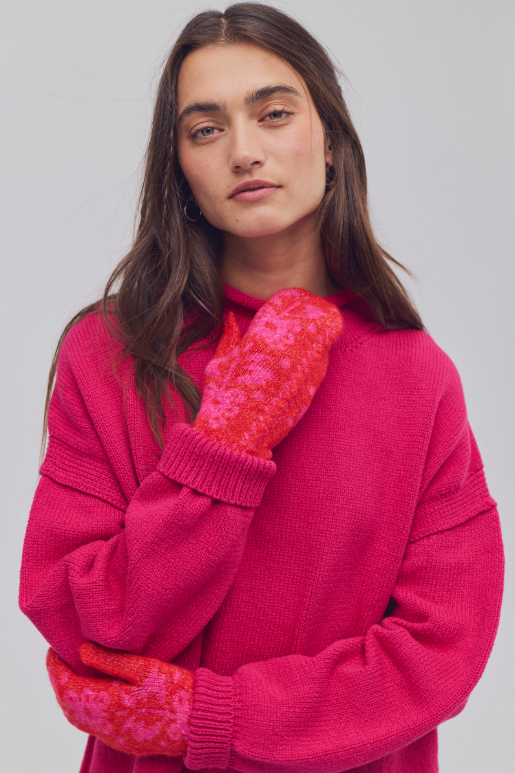 A woman wearing a bright pink knit sweater and red-orange knit mittens with a lighter pink floral pattern, posed against a light gray studio background.