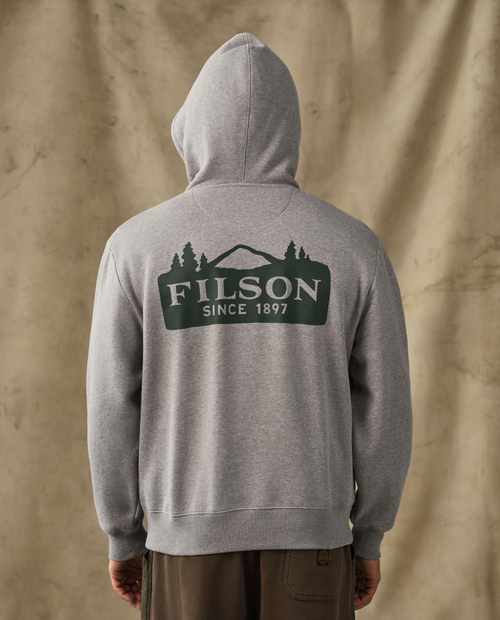 A person shown from the back wearing a heather gray pullover hoodie with a large dark green FILSON logo graphic on the back, paired with brown pants, set against a textured beige backdrop.