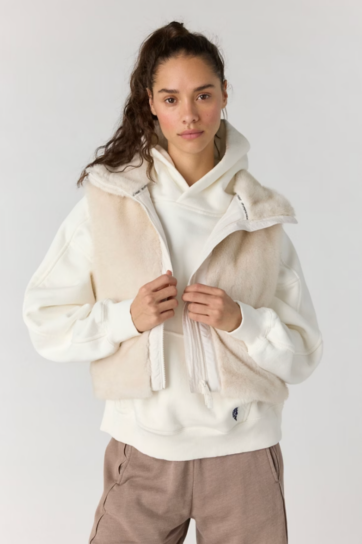 A model wearing a cream-colored hooded sweatshirt layered with a beige faux-shearling zip-up vest, paired with taupe pants, against a plain light gray studio background.