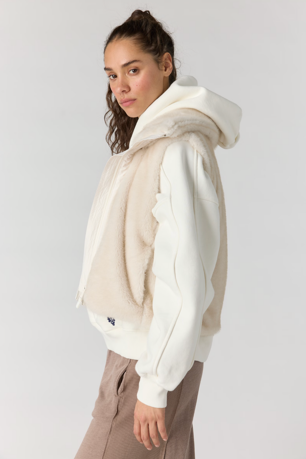 A woman wearing a cream-colored hooded jacket with a fuzzy beige fleece insert, paired with brown pants, photographed against a light gray studio background.