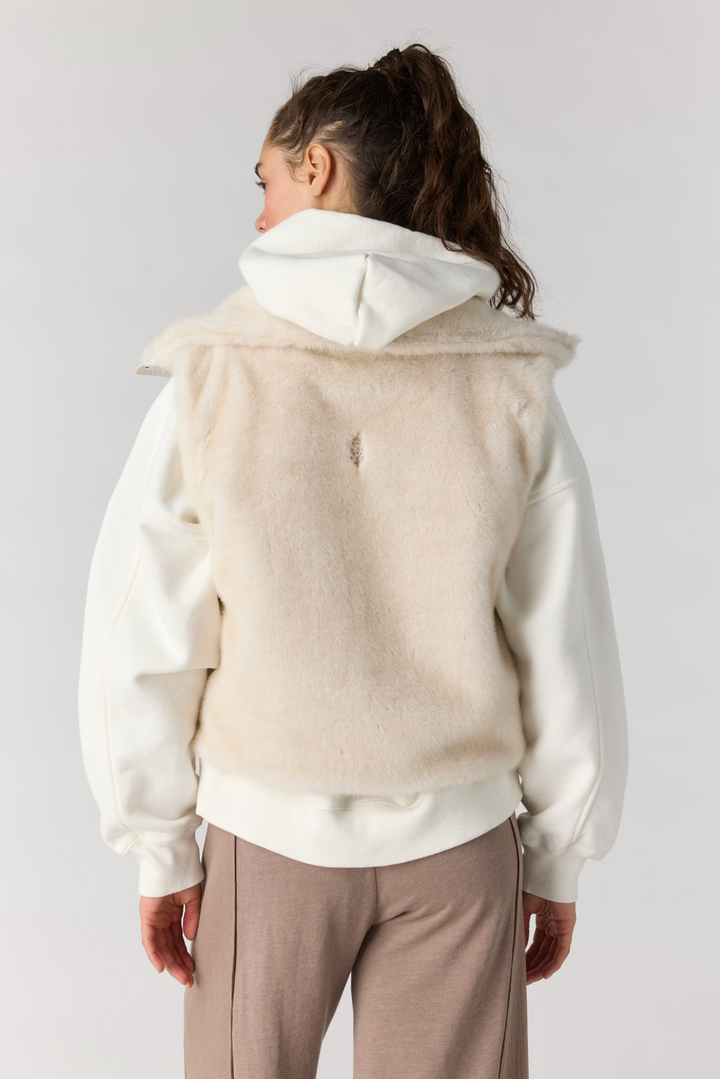 A model shown from the back wearing a cream-colored fuzzy hooded vest layered over a white sweatshirt and beige pants, set against a plain light gray studio background.