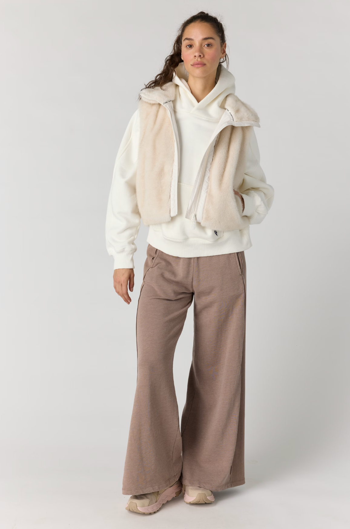 A model stands against a light gray studio background wearing a cream zip-front hoodie layered with a cream faux-fur vest, paired with wide-leg taupe pants and beige sneakers.