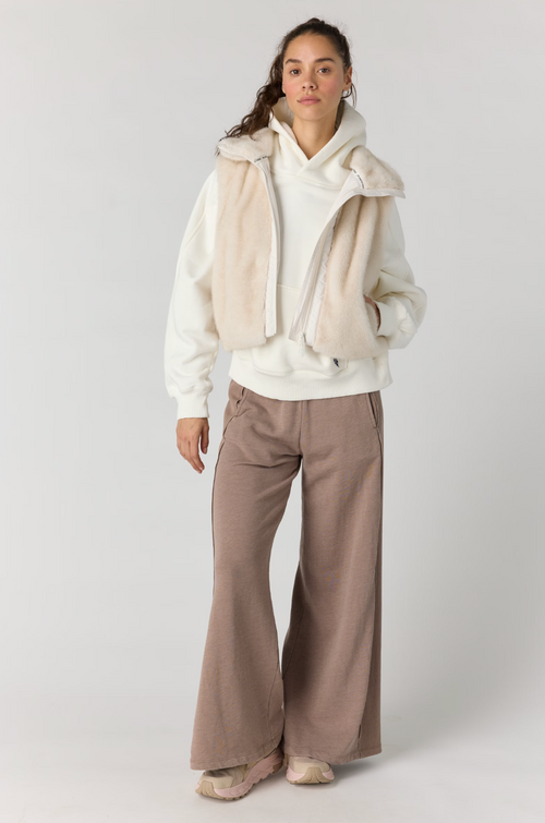 A model stands against a light gray studio background wearing a cream zip-front hoodie layered with a cream faux-fur vest, paired with wide-leg taupe pants and beige sneakers.
