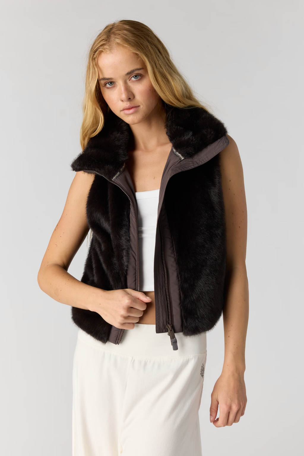 A blonde model wearing a sleeveless dark brown faux fur vest with a zippered front over a white top and white pants, photographed against a light gray studio background.