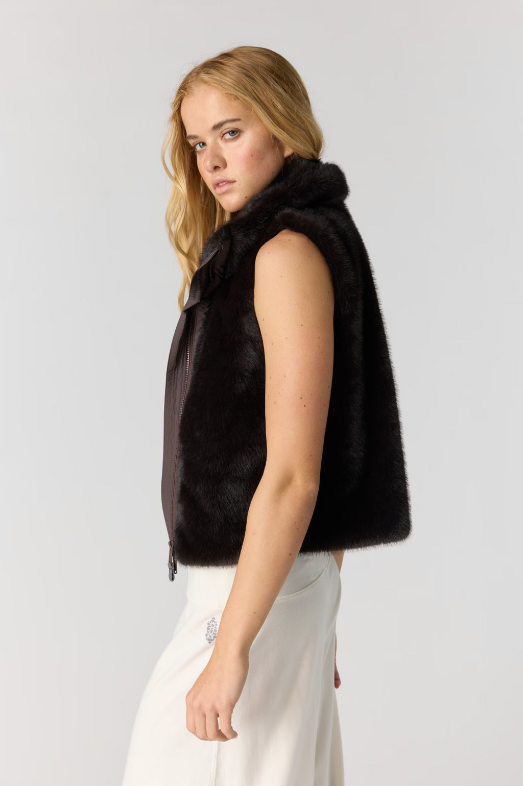 A woman shown in side profile wearing a sleeveless dark brown fur vest over a white skirt, photographed against a plain light gray studio background.