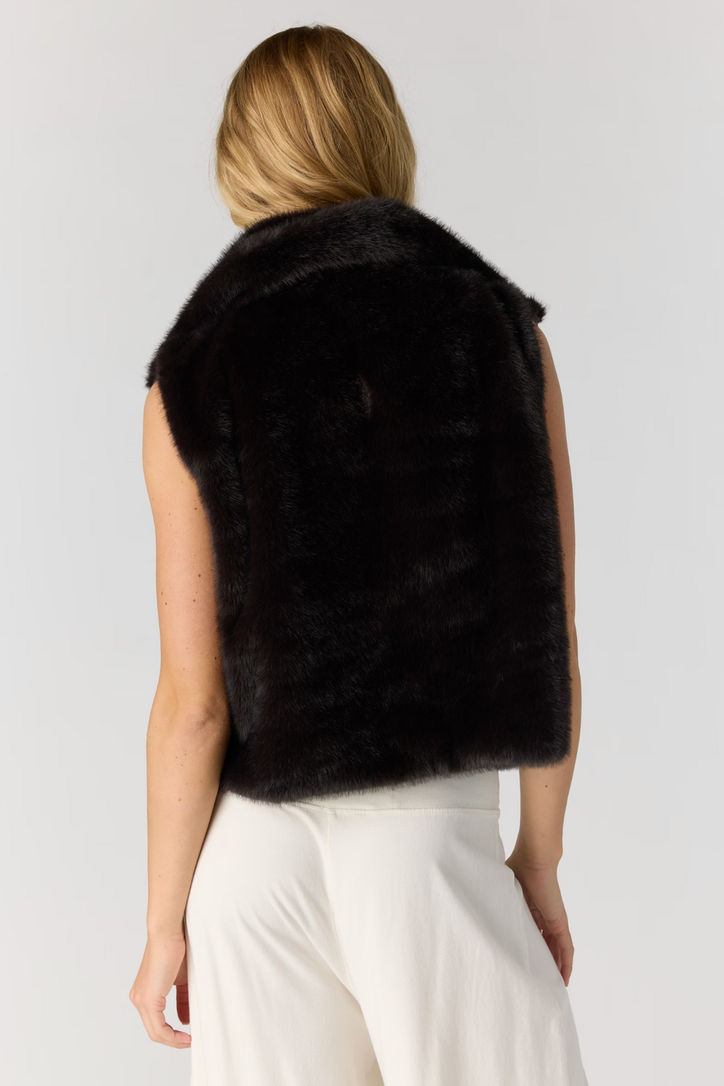A woman shown from the back wearing a sleeveless black faux fur vest over a white top and white pants, against a light gray studio background.