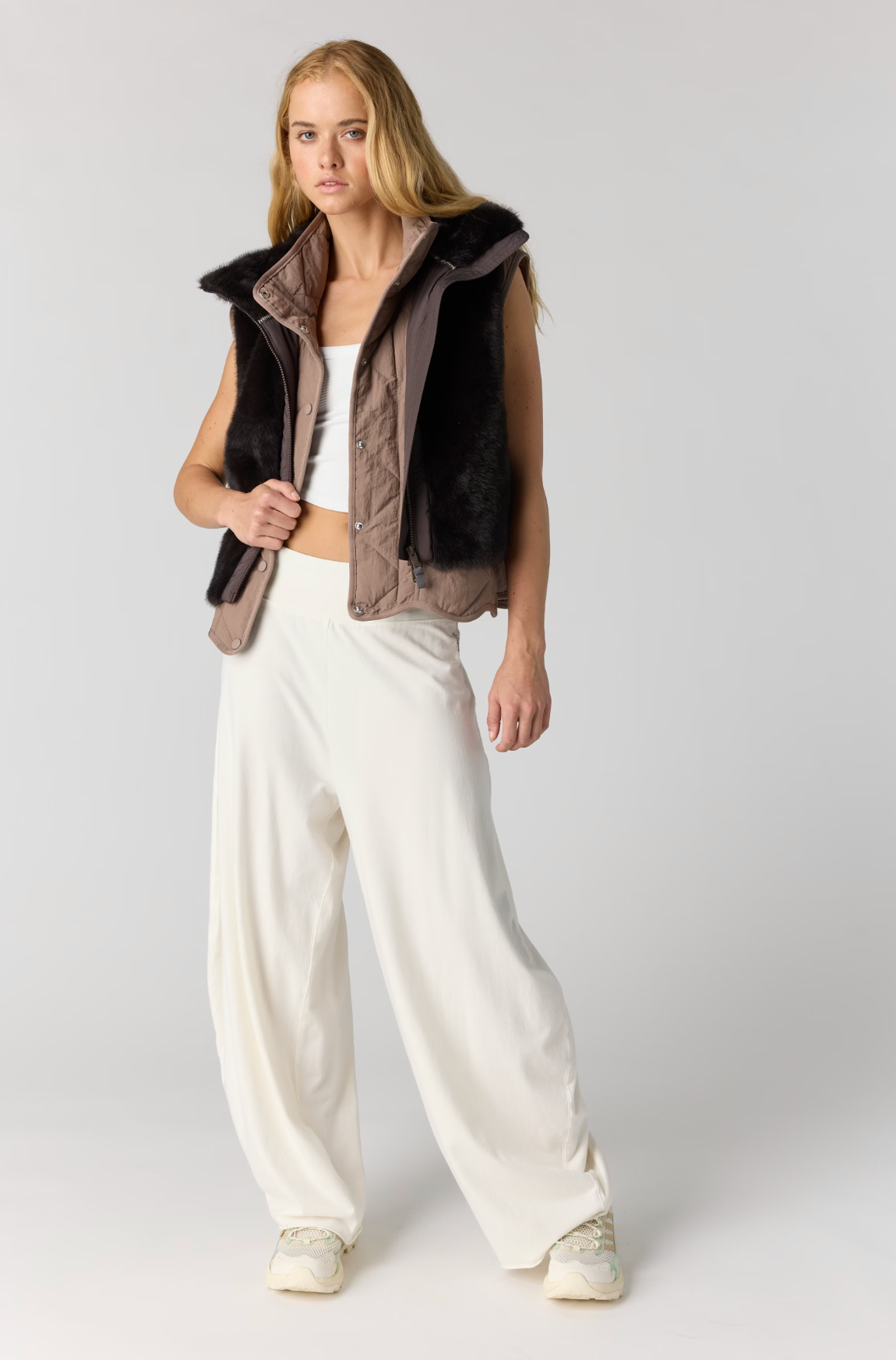 A model stands against a light gray studio background wearing a sleeveless quilted vest with dark brown faux-fur trim and a taupe-brown lining, styled over a white top and cream wide-leg trousers.