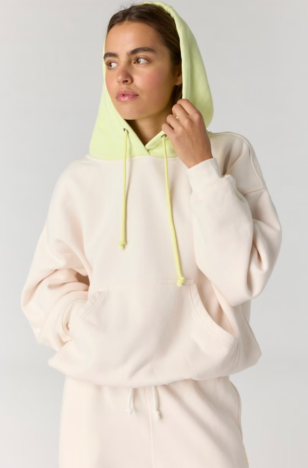A woman wearing a light cream-colored hoodie with a contrasting light green hood and drawstrings, posed against a neutral light gray background.