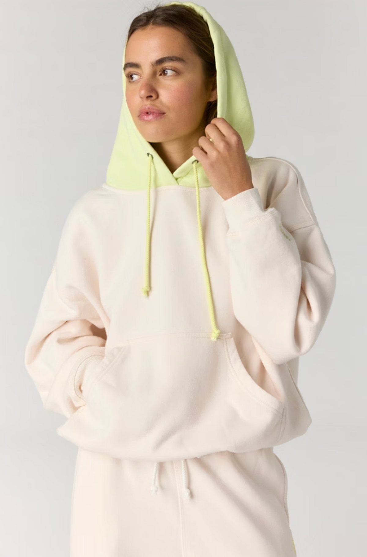 A woman wearing a light cream-colored hoodie with a contrasting light green hood and drawstrings, posed against a neutral light gray background.