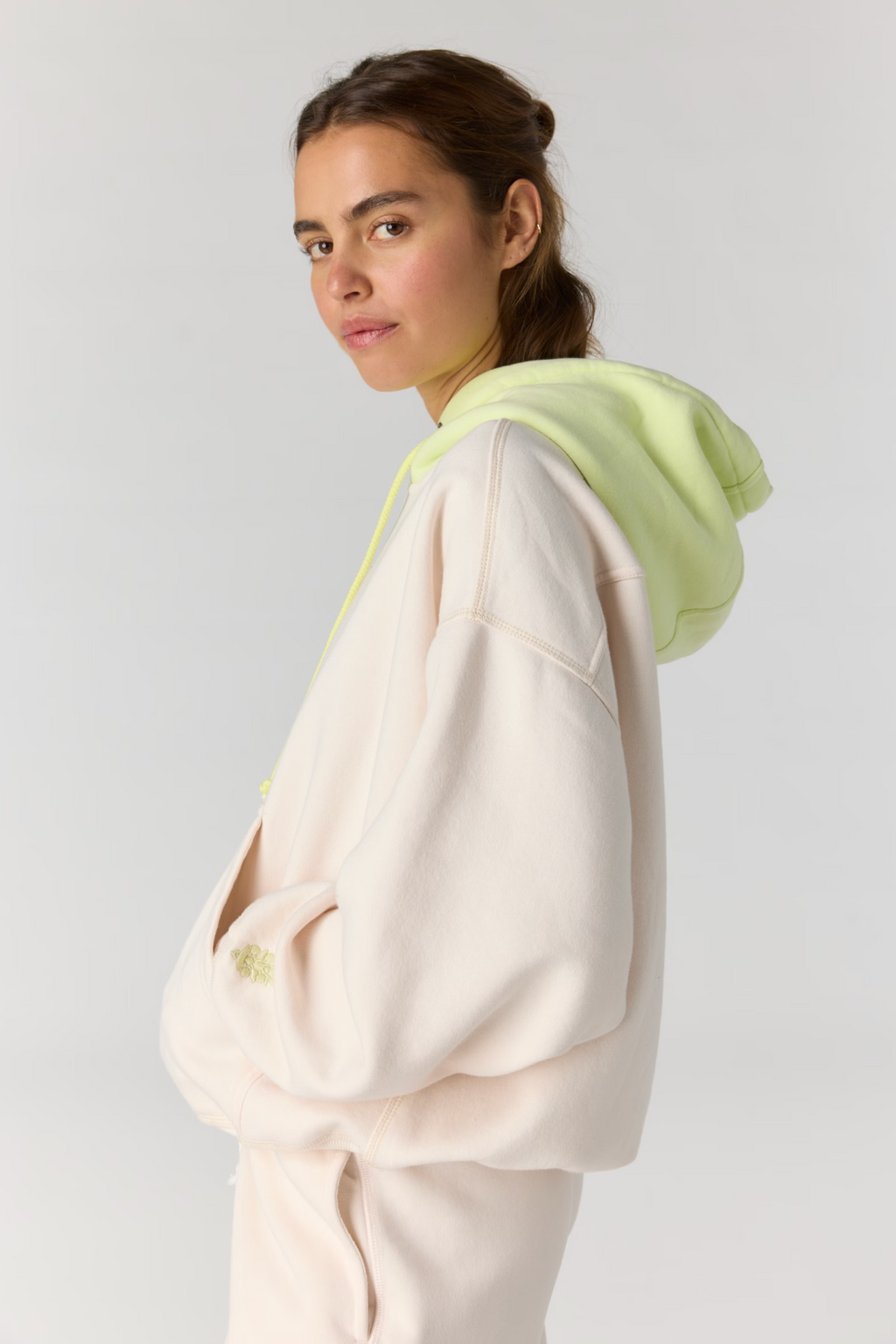 A woman wearing an oversized light cream hoodie with a bright neon yellow-green hood lining and drawstrings, photographed against a plain light gray studio background.