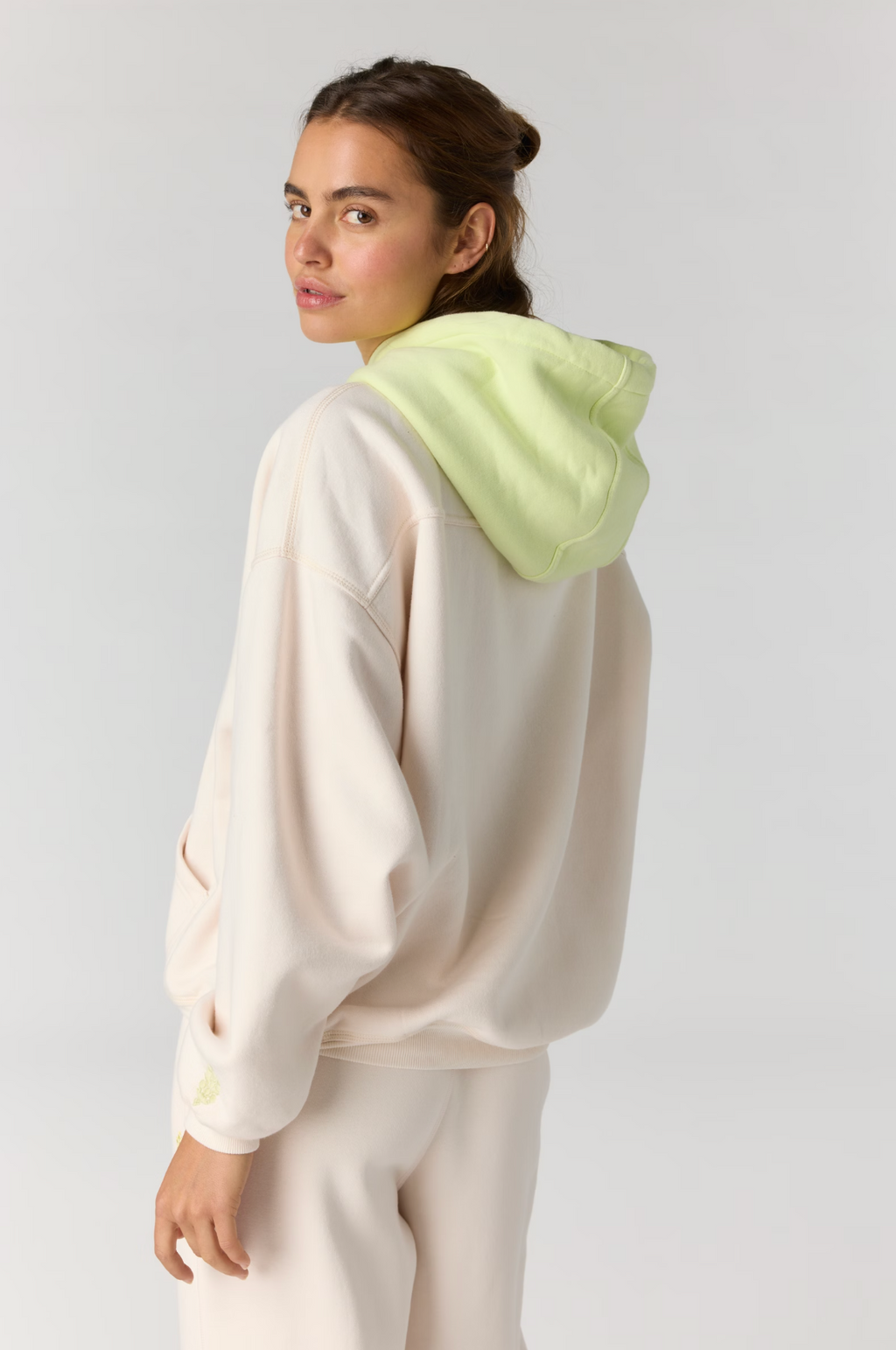 A woman wearing a cream-colored hoodie with a light green hood, photographed from the back against a light gray studio background.