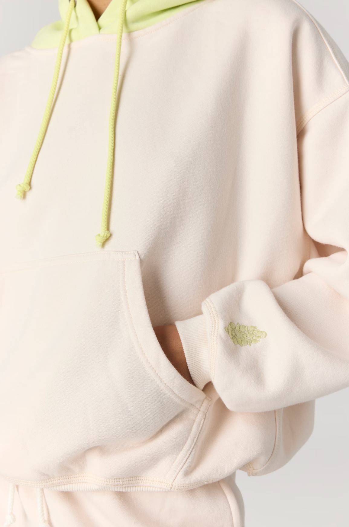 Close-up of a person wearing a cream-colored hoodie with light green drawstrings and a small light green embroidered leaf on the cuff.