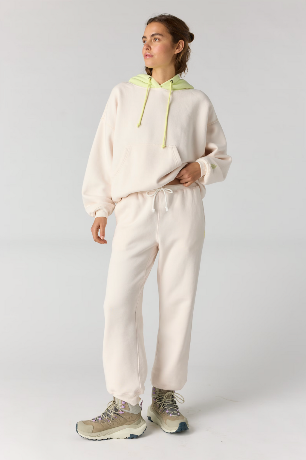 A woman modeling a matching cream-colored loungewear set consisting of a hoodie with a light green hood and drawstrings, and matching sweatpants, paired with beige sneakers against a light gray studio background.