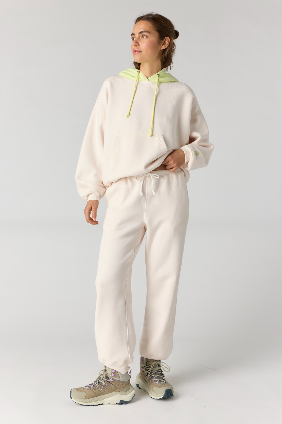 A woman modeling a matching cream-colored loungewear set consisting of a hoodie with a light green hood and drawstrings, and matching sweatpants, paired with beige sneakers against a light gray studio background.