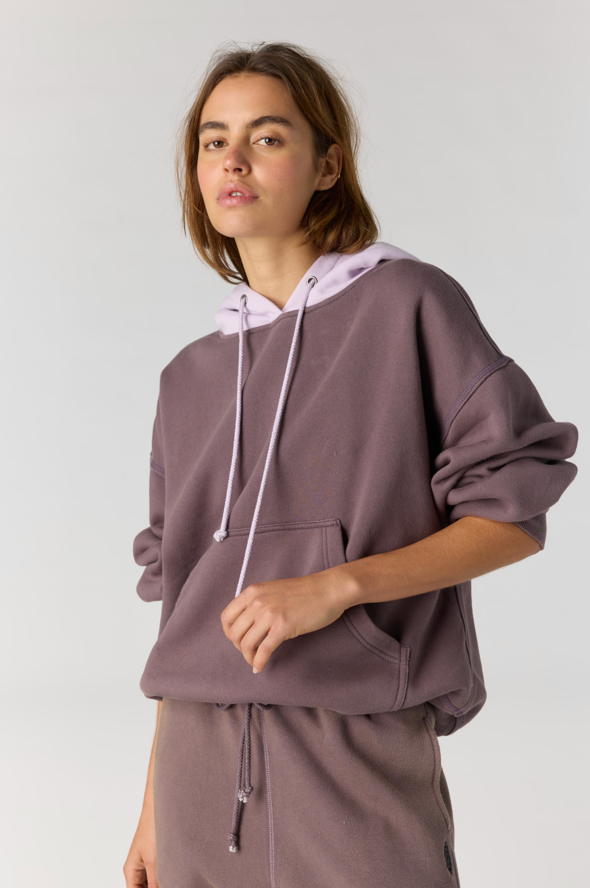 A woman modeling an oversized dusty mauve hoodie with a light lavender hood and drawstrings, paired with matching sweatpants, against a plain light gray studio background.