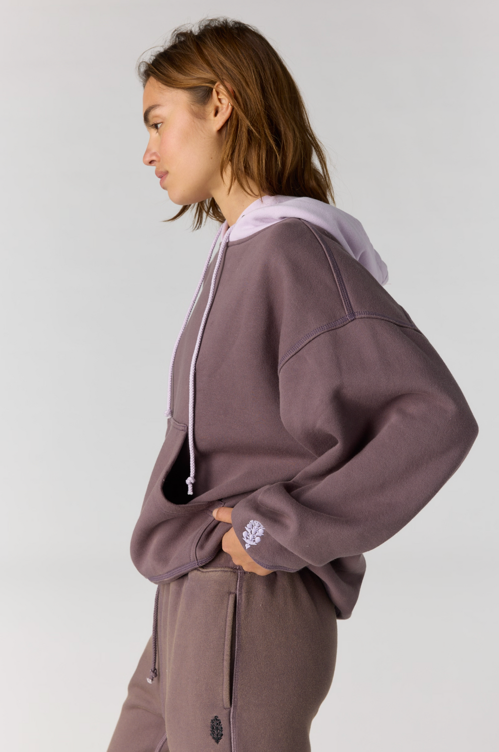 A woman in profile wearing a cropped, oversized hoodie and matching sweatpants in a dusty mauve-purple color, with a contrasting light lavender hood. The hoodie features white drawstrings and a small white embroidered logo on the cuff.