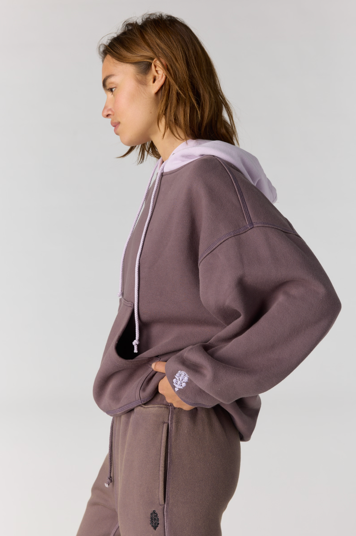 A woman in profile wearing a cropped, oversized hoodie and matching sweatpants in a dusty mauve-purple color, with a contrasting light lavender hood. The hoodie features white drawstrings and a small white embroidered logo on the cuff.