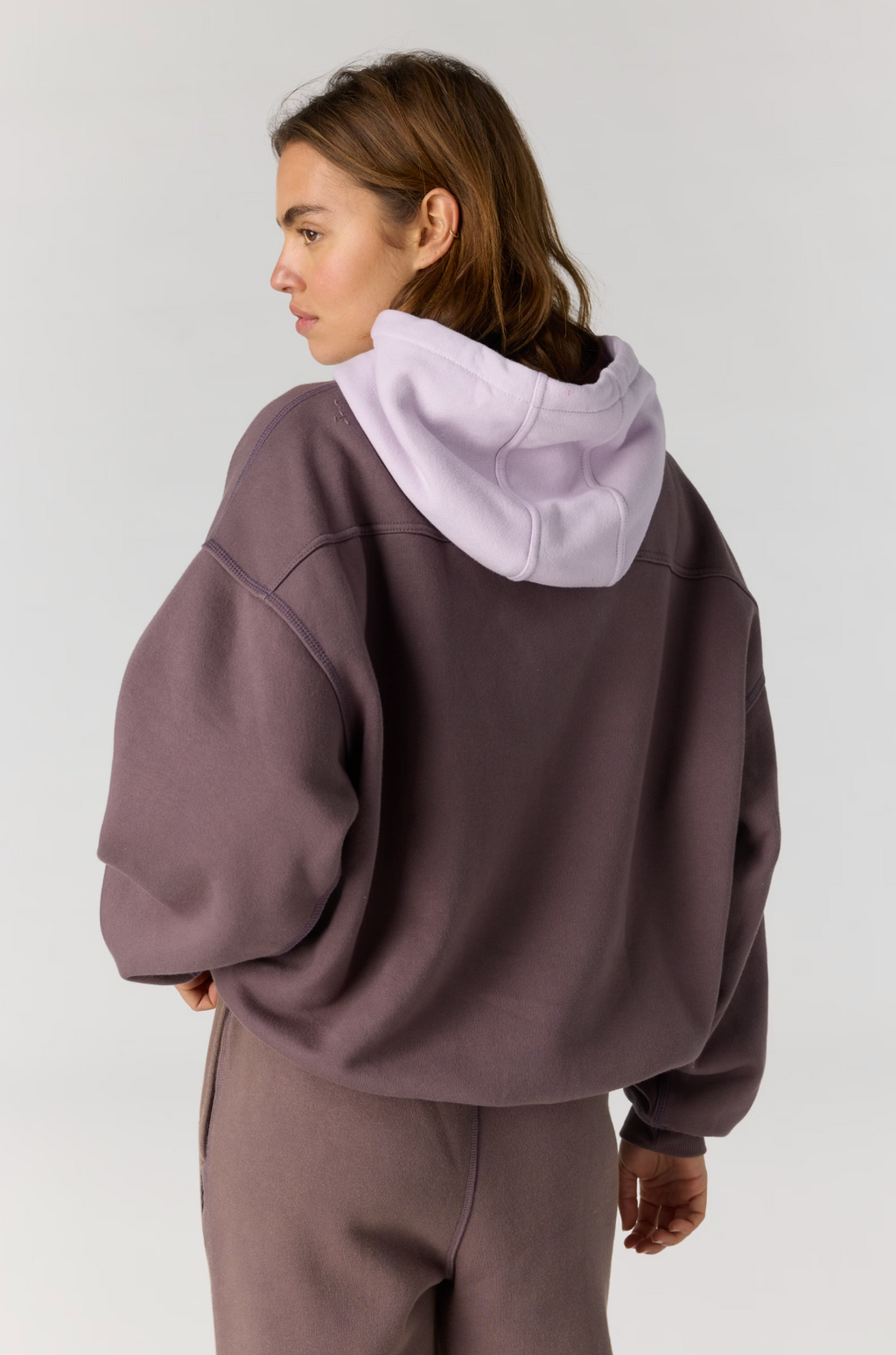 A woman shown from the back wearing an oversized mauve-brown hoodie with a contrasting light lavender hood, paired with matching sweatpants, against a plain light gray studio background.