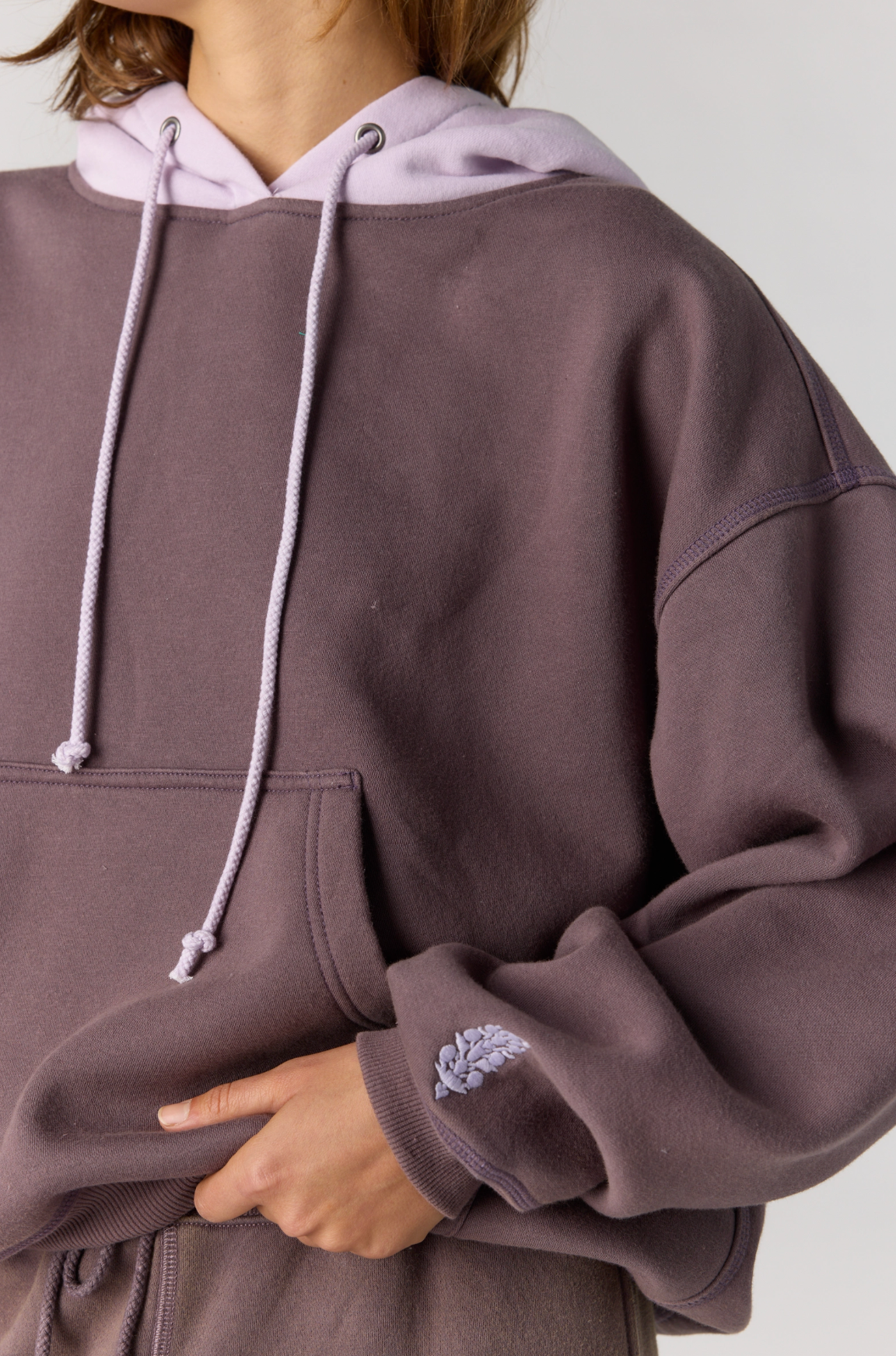 A close-up of a person wearing an oversized dusty mauve-purple hoodie with a light lavender hood lining and drawstrings, plus a small embroidered leaf detail on the cuff.