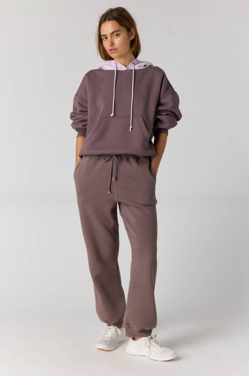 A woman modeling a matching two-piece loungewear set consisting of a cropped hoodie and sweatpants in a dusty mauve color, paired with white sneakers, against a light gray studio background.