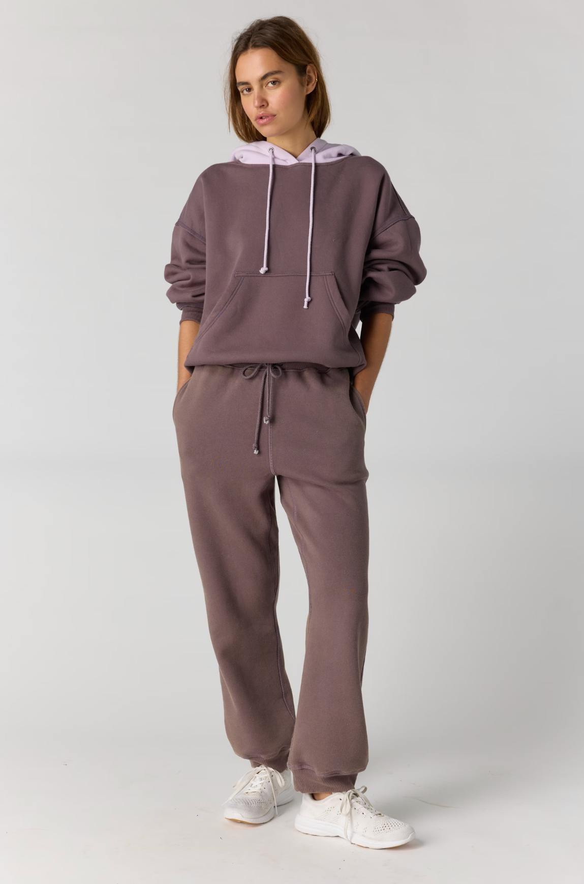 A woman modeling a matching two-piece loungewear set consisting of a cropped hoodie and sweatpants in a dusty mauve color, paired with white sneakers, against a light gray studio background.