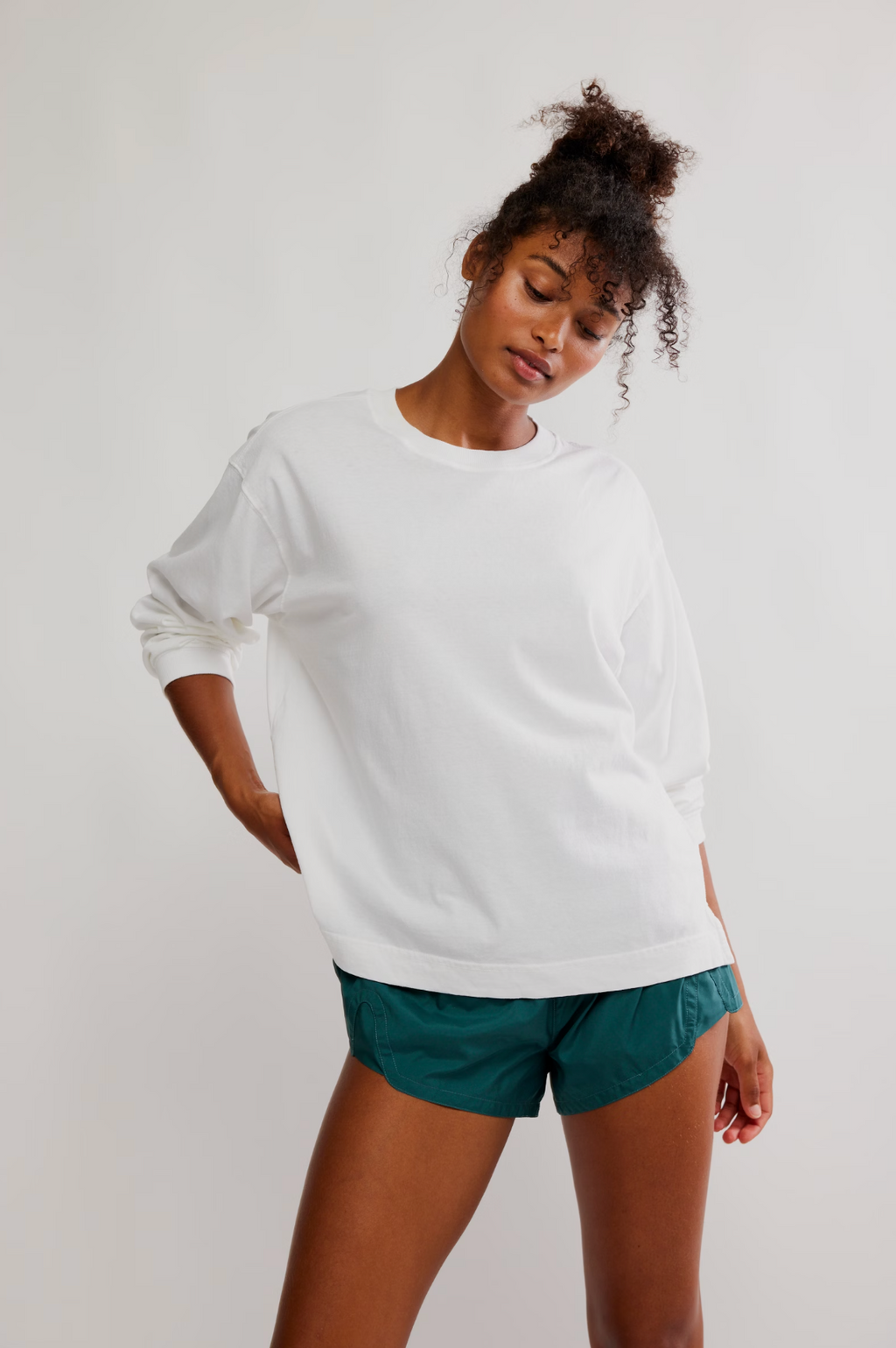 A woman wearing a white long-sleeve crewneck sweatshirt and teal athletic shorts, posing against a light gray studio background.