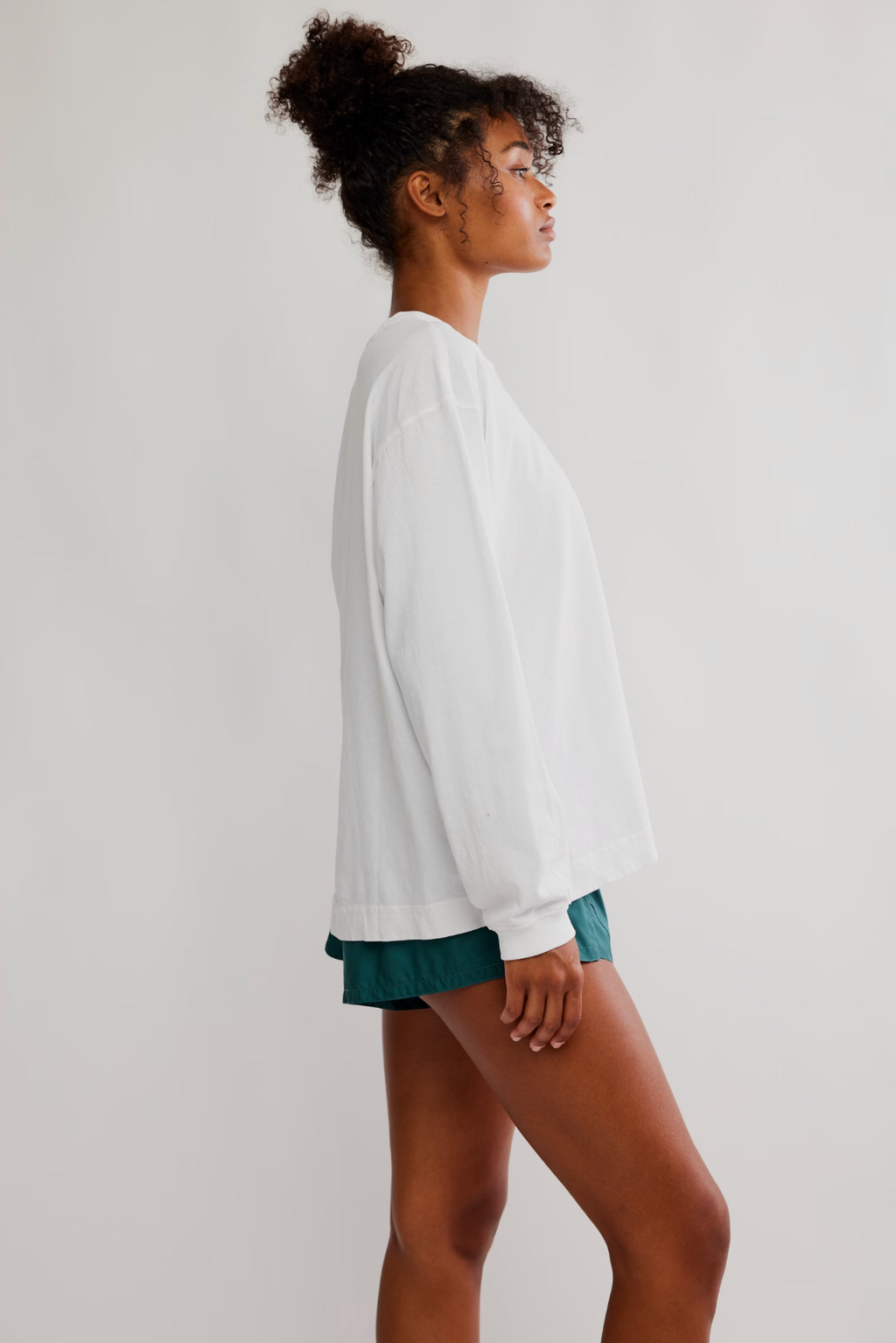 A woman in side profile wearing a white long-sleeve crewneck sweatshirt and teal shorts, standing against a plain light gray studio background.