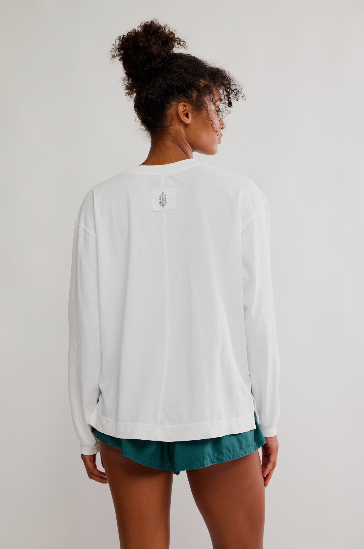 A woman shown from the back wearing a white long-sleeve crewneck shirt with a small graphic on the upper back and teal shorts, set against a plain light gray studio background.