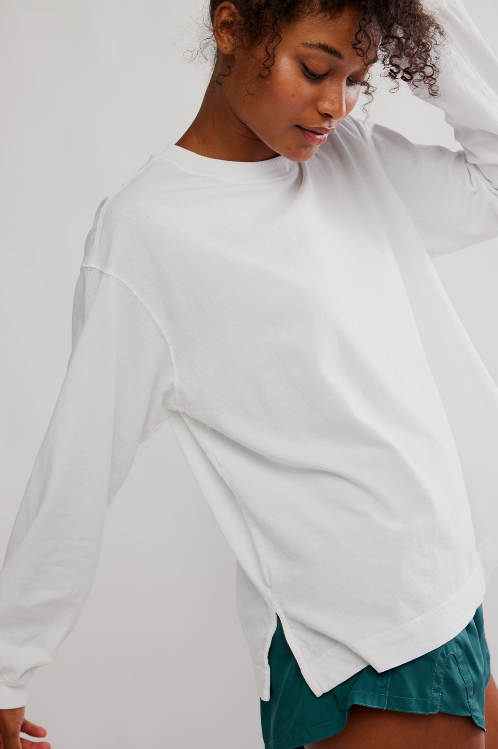A woman wearing a white long-sleeve crewneck shirt and teal shorts, captured in a close-up shot against a plain light background.