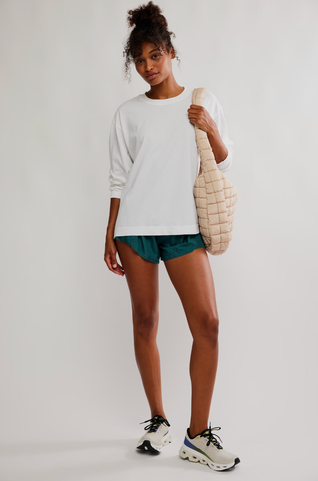 A woman modeling a white long-sleeve crewneck sweatshirt, teal athletic shorts, and white sneakers, carrying a beige quilted shoulder bag against a plain light gray studio background.