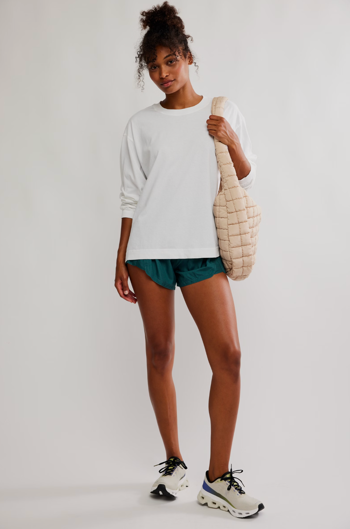 A woman modeling a white long-sleeve crewneck sweatshirt, teal athletic shorts, and white sneakers, carrying a beige quilted shoulder bag against a plain light gray studio background.