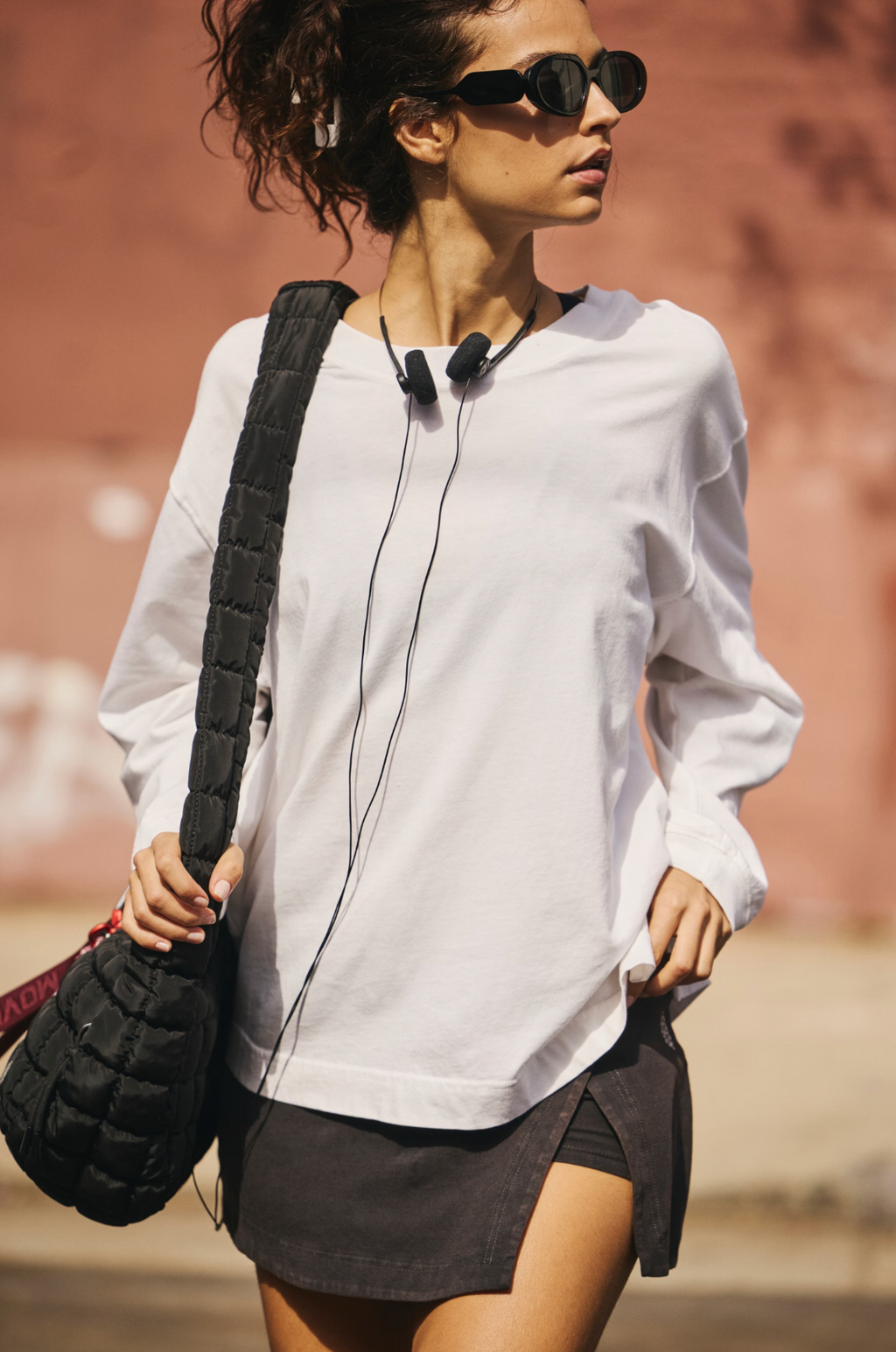 A woman wearing a white long-sleeve crewneck sweatshirt, a dark gray wrap-style mini skirt, black sunglasses, and carrying a black quilted shoulder bag, walking outdoors against a warm terracotta wall.