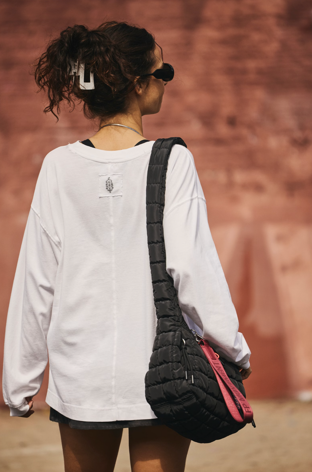 A woman from behind wearing a white oversized long-sleeve shirt and sunglasses, carrying a black quilted shoulder bag with a pink accent strap against a warm reddish-brown wall background.