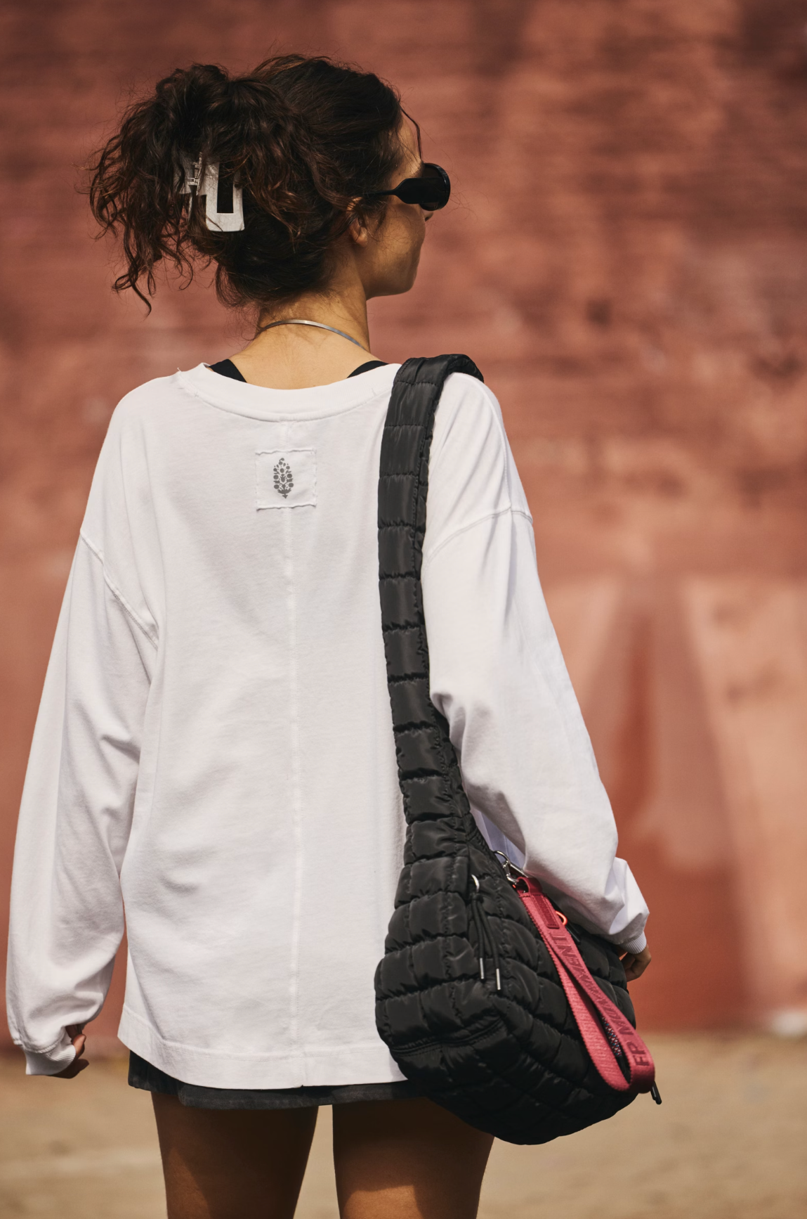 A woman from behind wearing a white oversized long-sleeve shirt and sunglasses, carrying a black quilted shoulder bag with a pink accent strap against a warm reddish-brown wall background.
