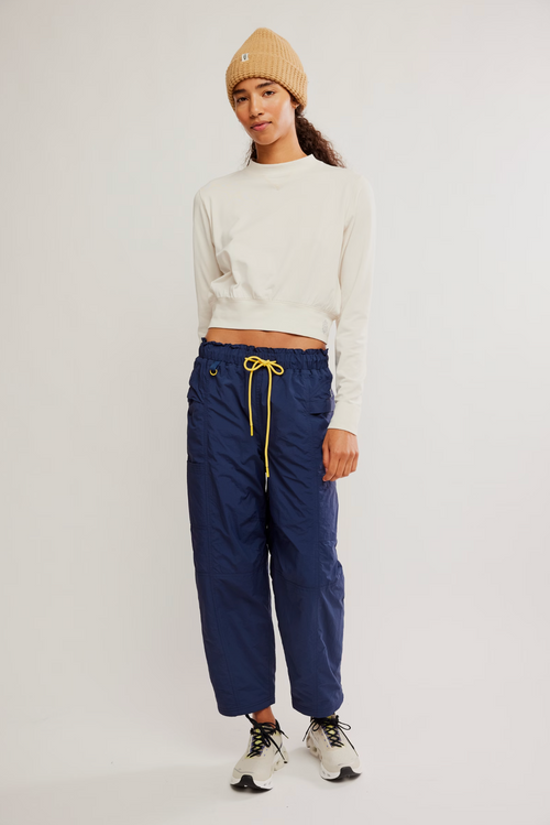 A model wearing navy blue drawstring pants, a white long-sleeve crop top, a beige knit beanie, and white sneakers, standing against a light gray studio background.