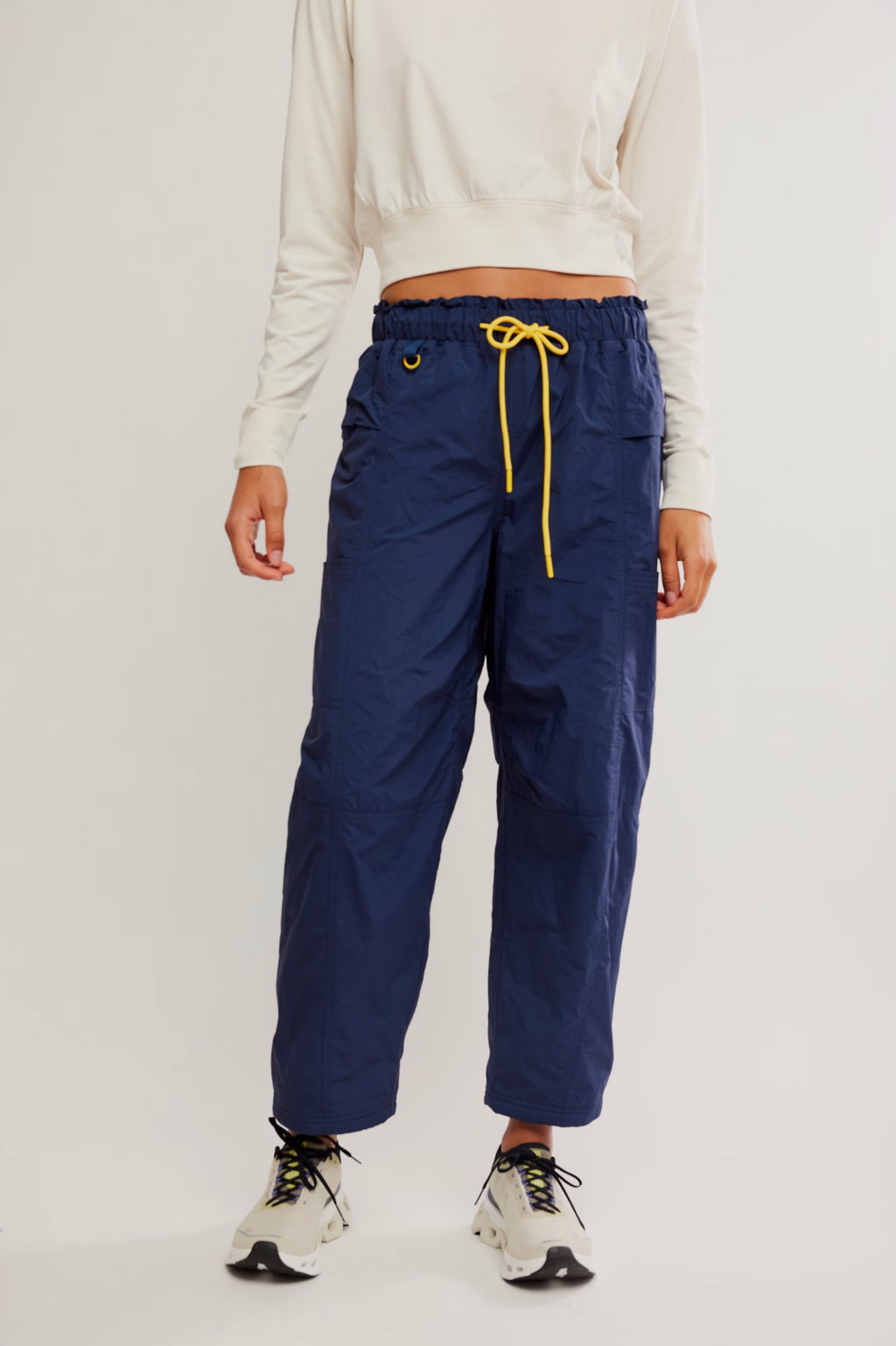 A model is wearing navy blue wide-leg cargo pants with a yellow drawstring, styled with a cream long-sleeve crop top and white sneakers against a plain light gray studio background.