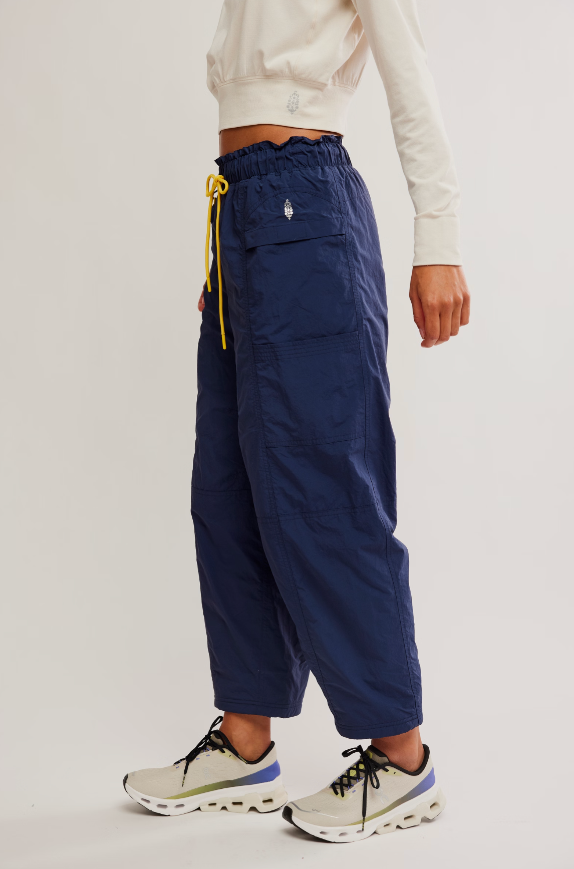 A person wearing navy blue wide-leg cargo pants with a yellow drawstring, styled with a cream cropped sweatshirt and light-colored sneakers, photographed against a plain light gray studio background.