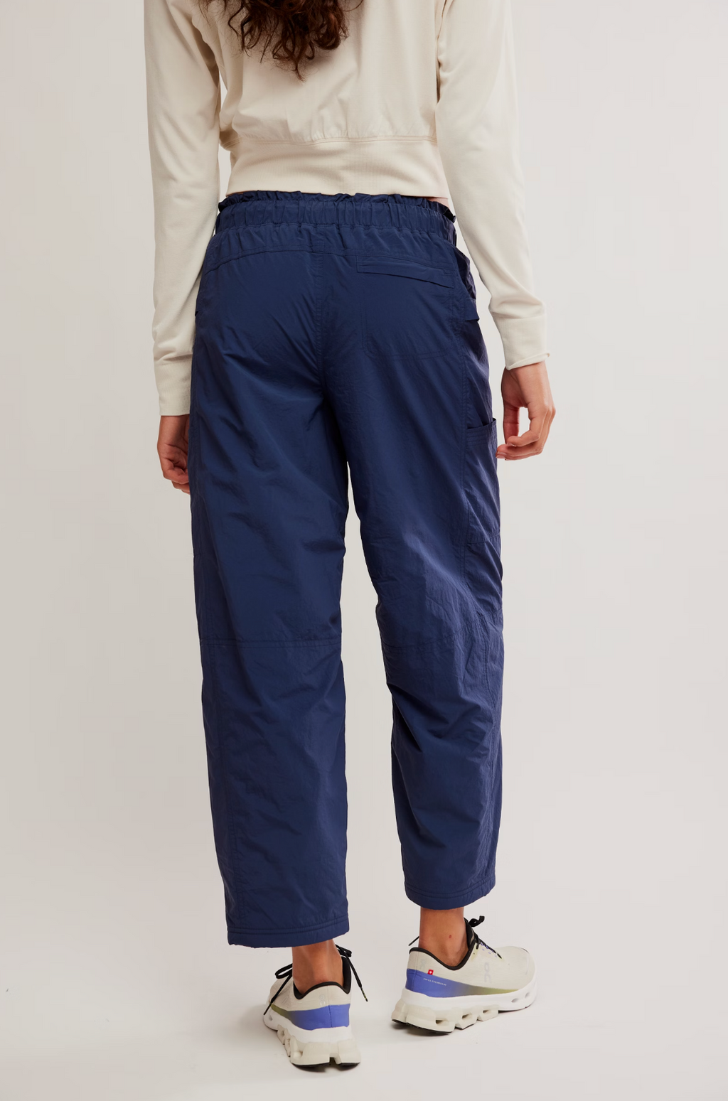 Rear view of a person wearing navy blue cropped cargo pants, a cream long-sleeve top, and white sneakers with blue accents, against a plain light background.