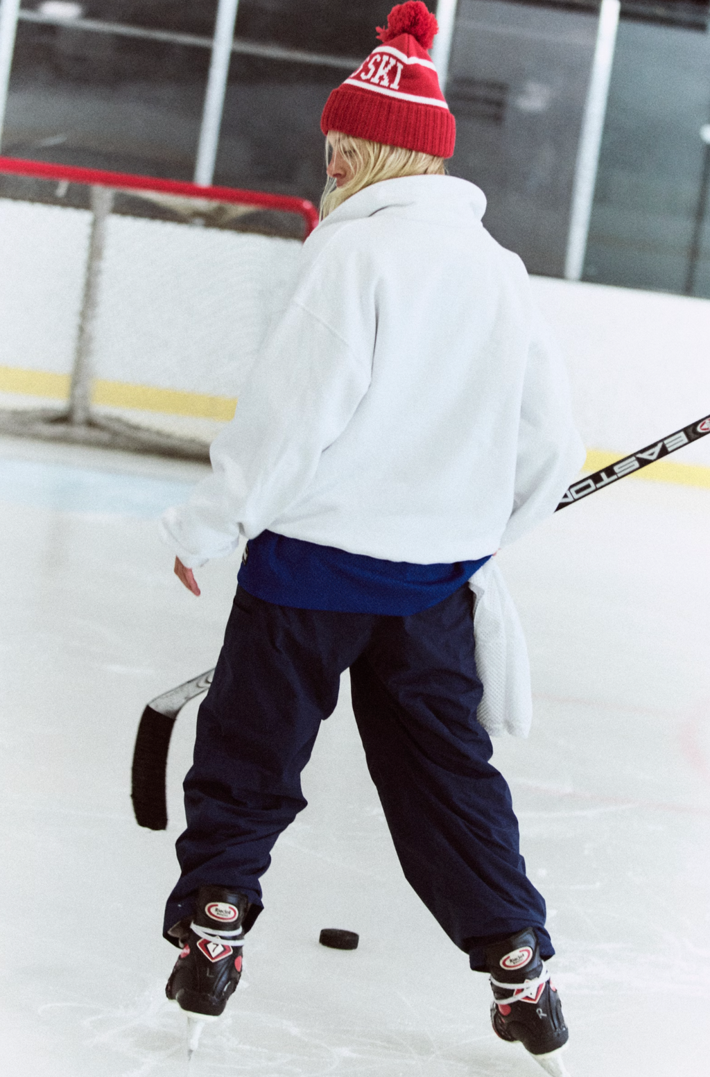 A person on an ice rink viewed from behind, wearing a white oversized hoodie, dark navy pants, a red knit beanie with white lettering, and hockey skates, holding a hockey stick.