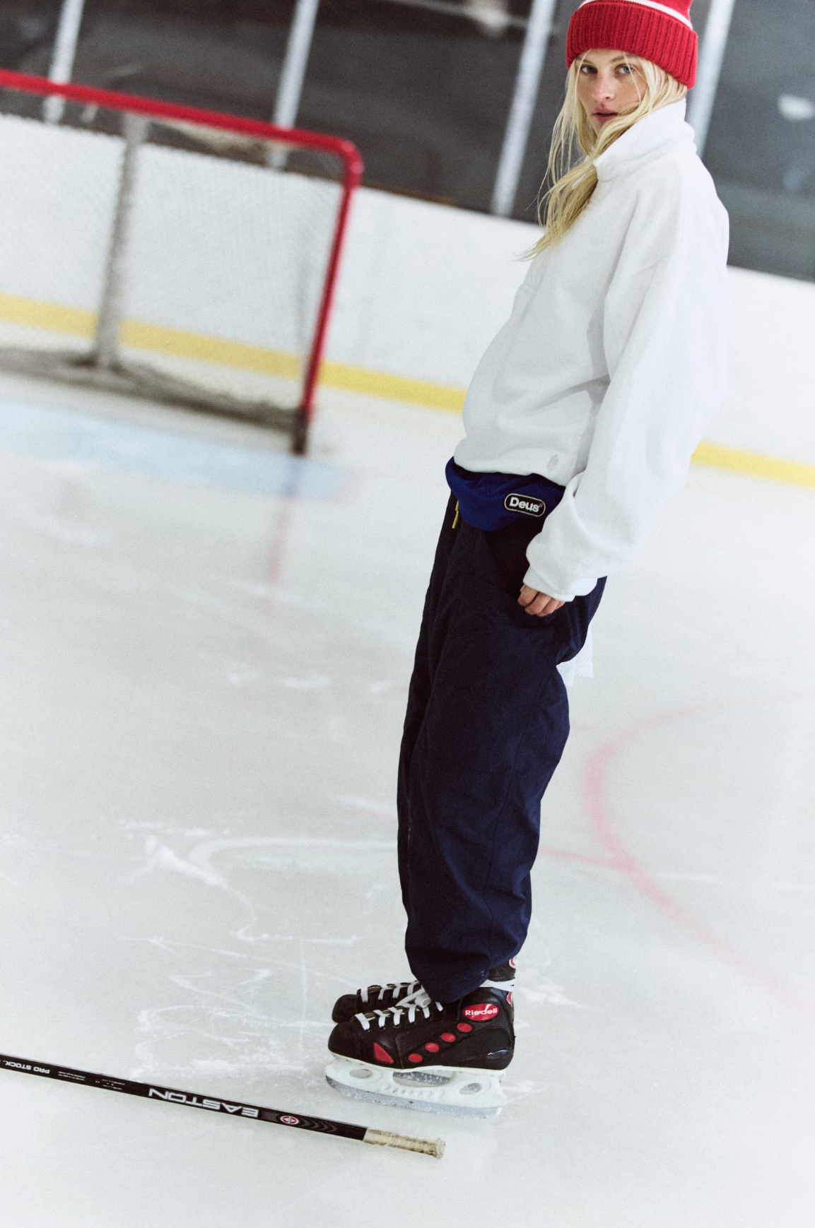 A person standing on an ice rink wearing a white sweatshirt, dark navy pants, a red beanie, and black hockey skates with red accents. A hockey stick is visible in the foreground, and an ice hockey goal is in the background.