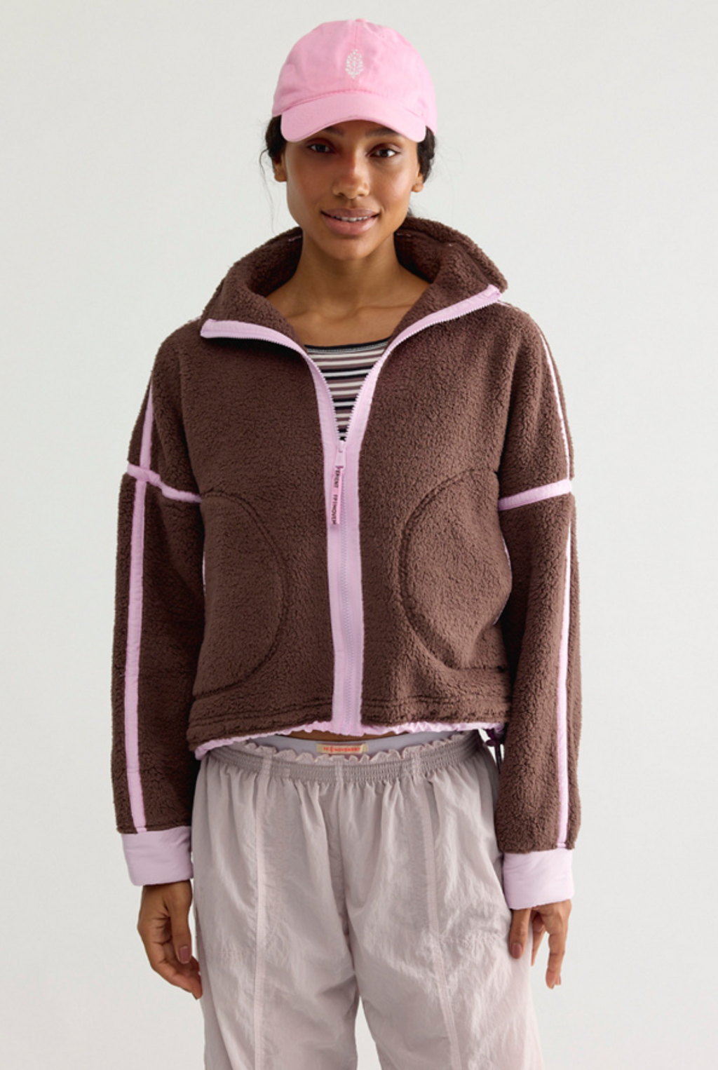 A model wearing a brown fleece zip-up jacket with light pink trim and stripes, a pink baseball cap, a striped shirt, and light gray sweatpants, photographed against a plain light gray studio background.