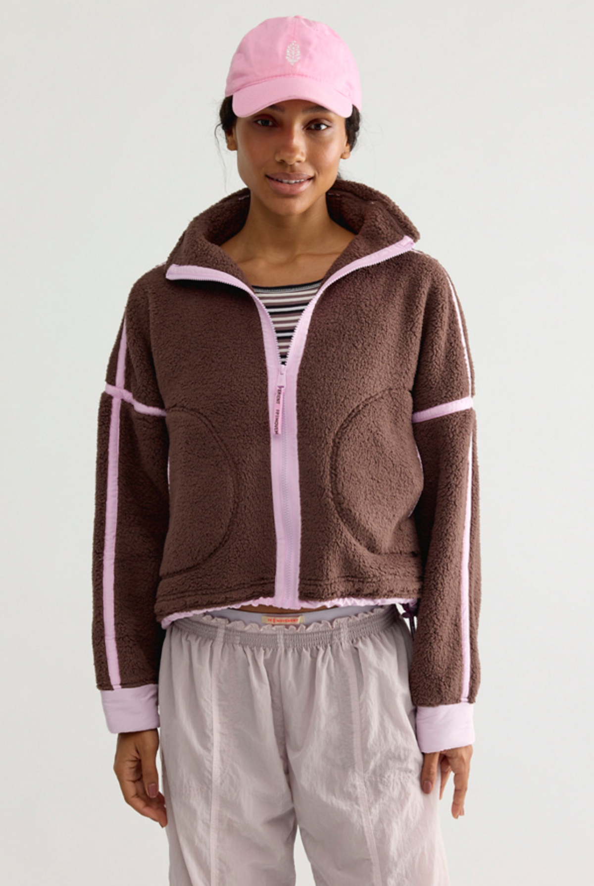 A model wearing a brown fleece zip-up jacket with light pink trim and stripes, a pink baseball cap, a striped shirt, and light gray sweatpants, photographed against a plain light gray studio background.