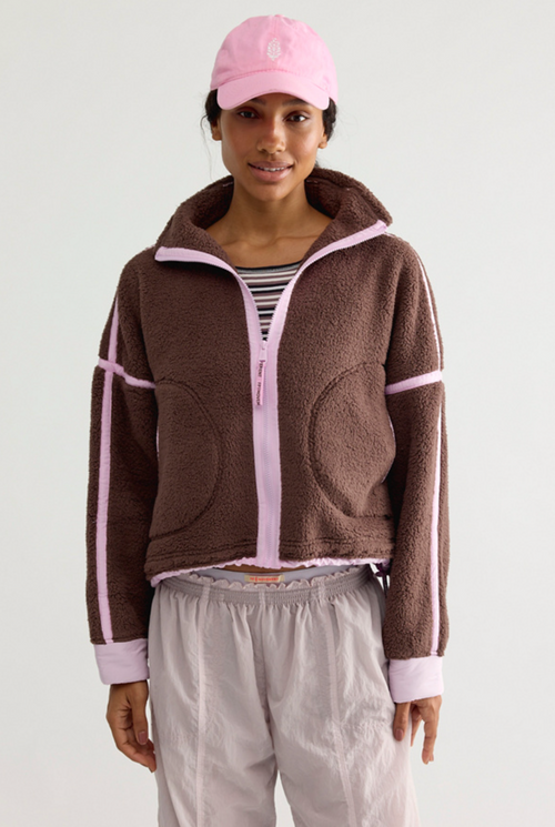 A model wearing a brown fleece zip-up jacket with light pink trim and stripes, a pink baseball cap, a striped shirt, and light gray sweatpants, photographed against a plain light gray studio background.