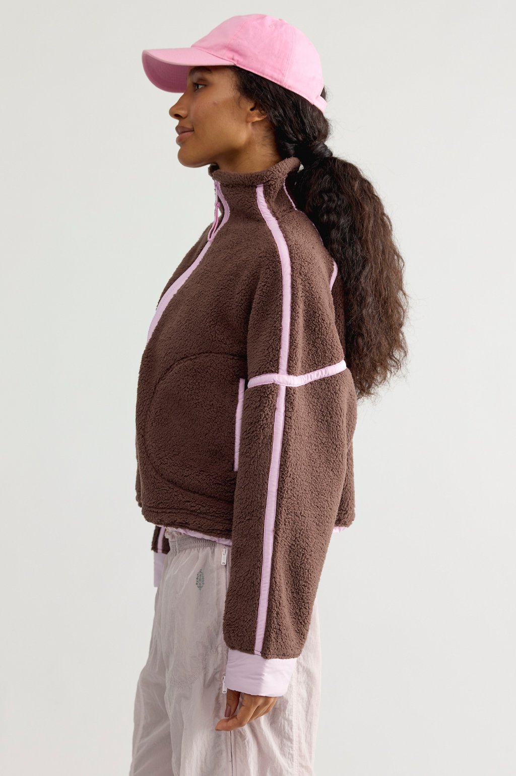 A woman in profile wearing a brown fleece zip-up jacket with light pink contrast piping, a pink baseball cap, and light pink pants, photographed against a plain light gray studio background.