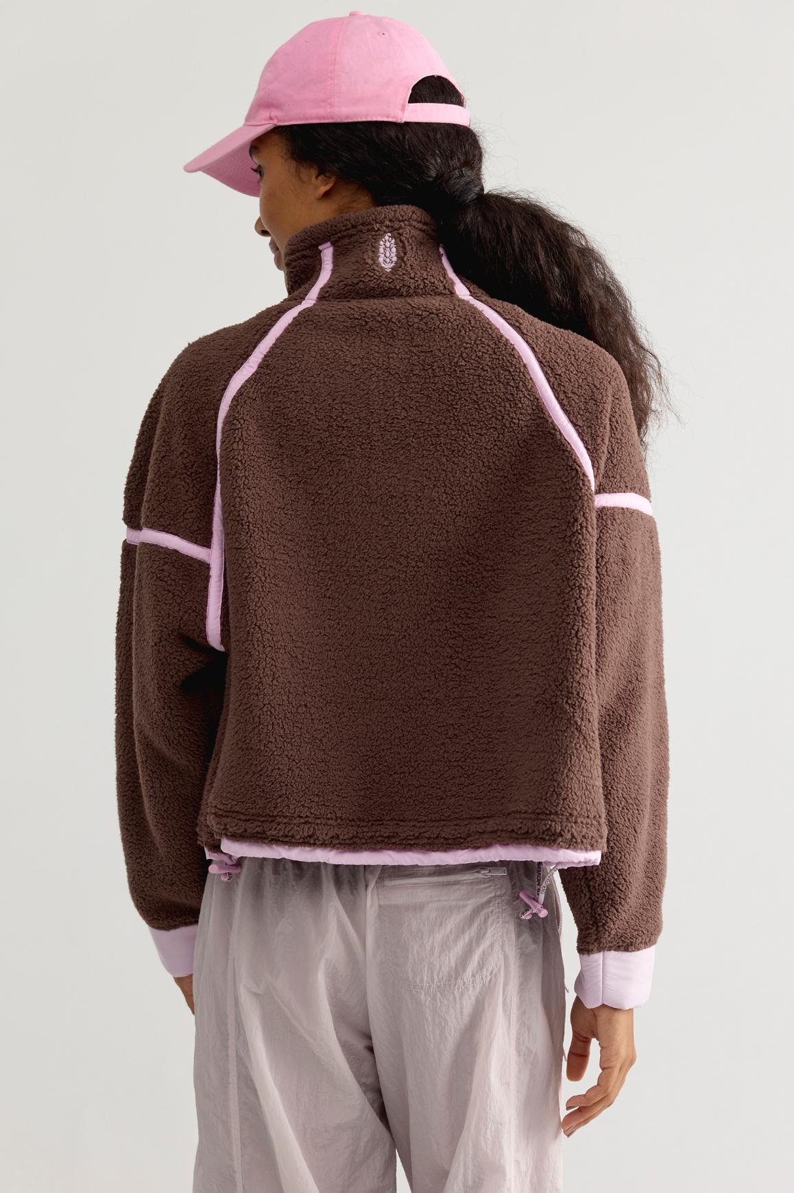 A person shown from the back wearing a brown sherpa fleece zip-up jacket with light pink contrast stitching and trim, a pink baseball cap, and light gray pants, against a plain light gray studio background.