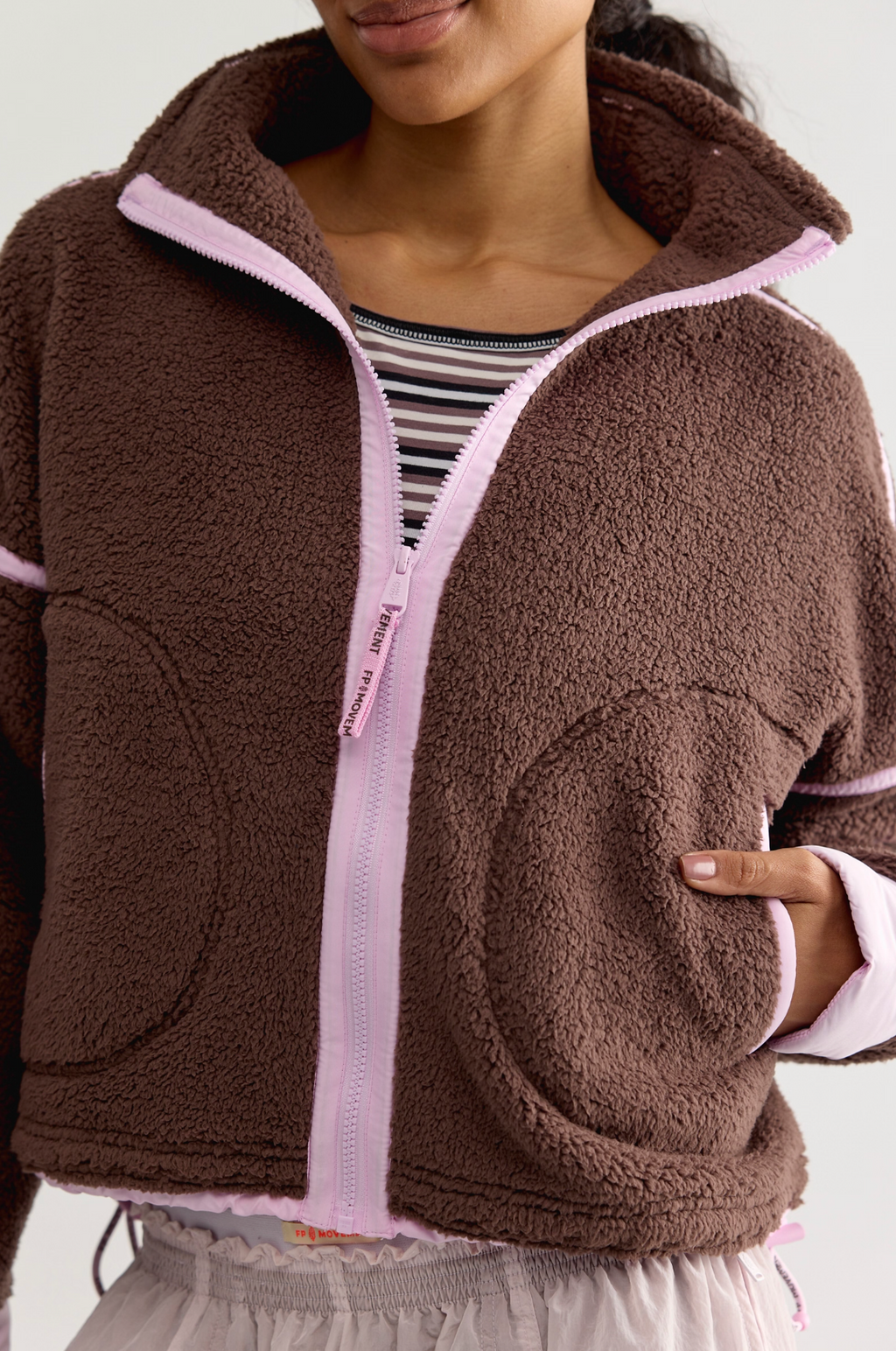 A close-up of a person wearing a brown sherpa fleece zip-up jacket with light pink trim and a striped shirt underneath, against a plain light background.