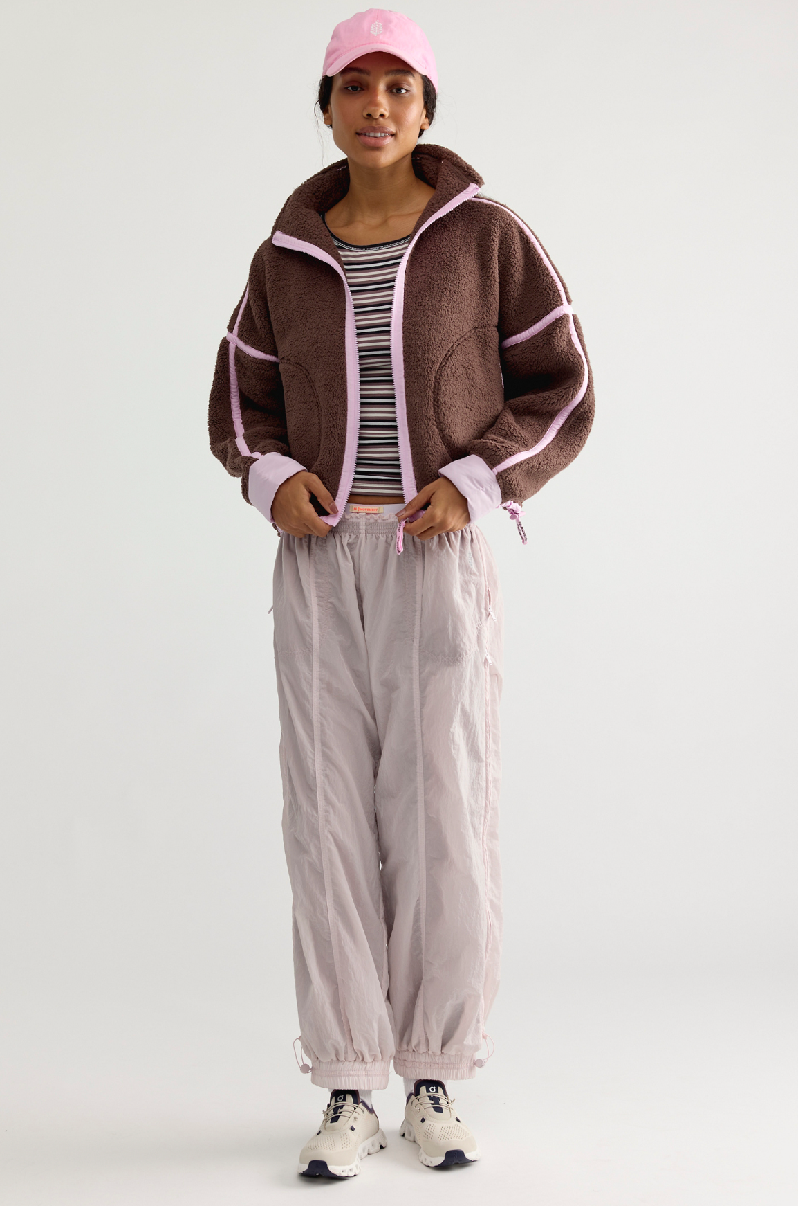 A model stands against a light gray studio background wearing a brown sherpa zip-up jacket with pink trim, light gray jogger pants, a black and white striped t-shirt, a pink baseball cap, and white sneakers.